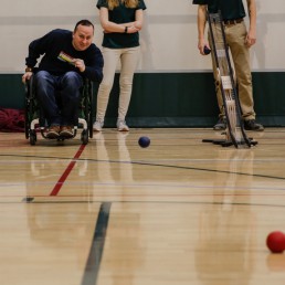Man in wheelchair enjoying game of bocce, throwing blue ball.